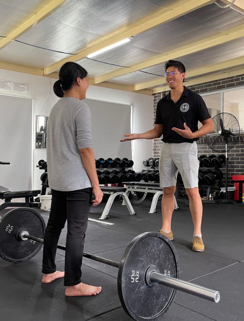 a Smiling Male Trainer in a Black Polo Shirt and Gray Shorts Gestures Instructively to a Barefoot Female Client in a Gray Top and Black Leggings As They Stand Beside a Loaded Barbell on the Floor of a Modern Gym Filled with Weights and Equipment   